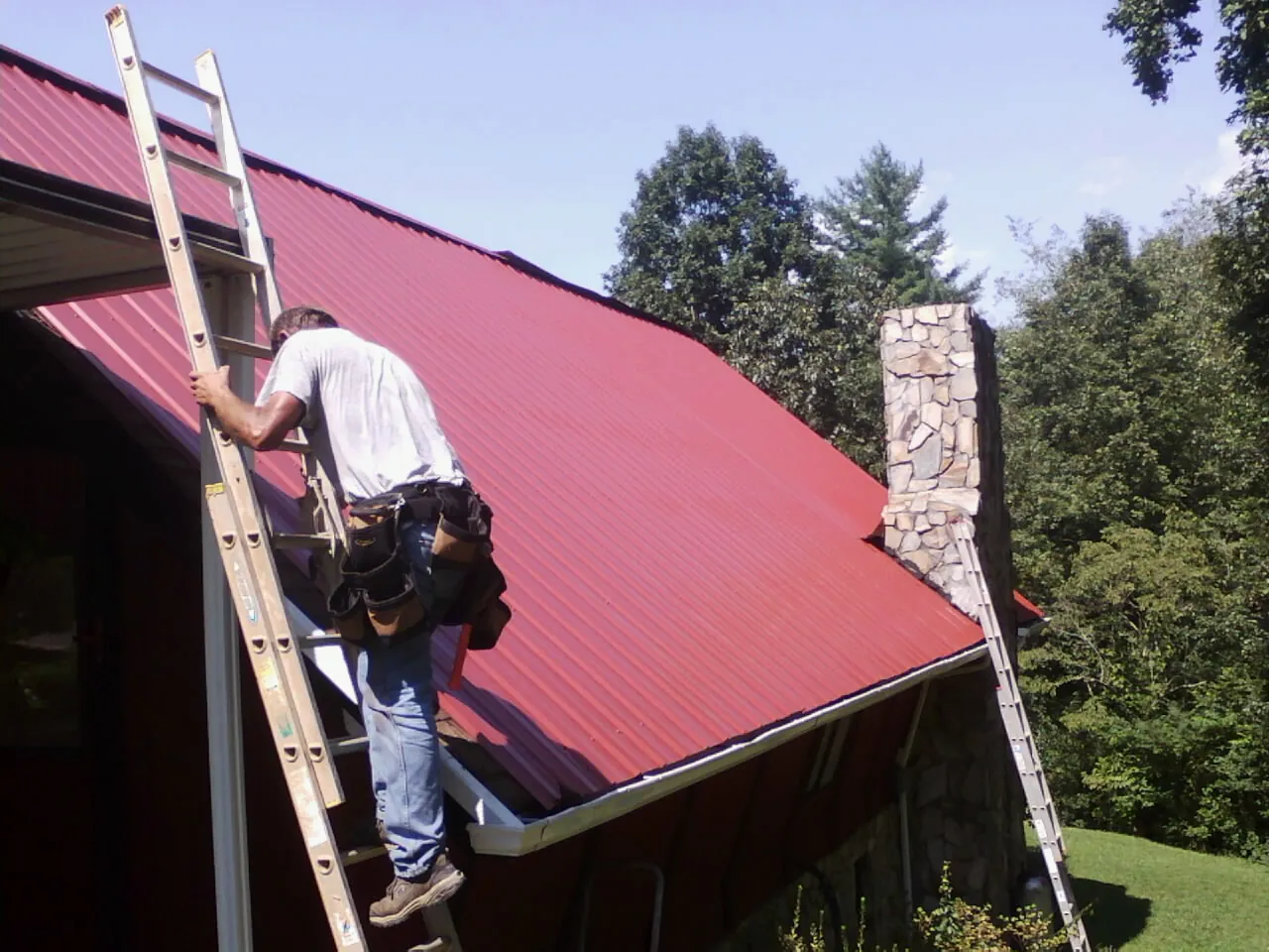 Roofer on a ladder beside a red metal roof and stone chimney — Soffit & Fascia Repair by Roofing Brothers in Chapel Hill